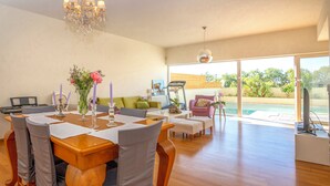 Un comedor amplio con una mesa de madera dispuesta para cuatro personas, rodeada de sillas con cojines. Un candelabro cuelga del techo, y un ventanal ofrece vistas a una piscina y follaje exterior.