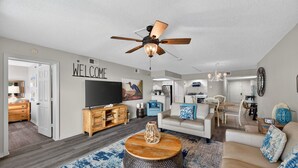 Un salon moderne avec un meuble de rangement en bois pour les appareils électroniques, un ventilateur de plafond et un panneau mural portant l’inscription « BIENVENUE ».