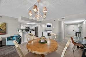 A modern kitchen with white cabinets, a microwave, and a refrigerator. There is a dining area with a wooden table and chairs. - Beachfront Bliss with Gulf Views, Balcony, Heated Pool & More (Indian Shores)