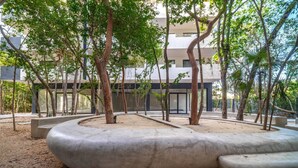 A landscaped courtyard with trees, a curved concrete bench, and a building in the background. - Tulum 2BR | Two Floors | Private Terrace + Grill (Tulum)
