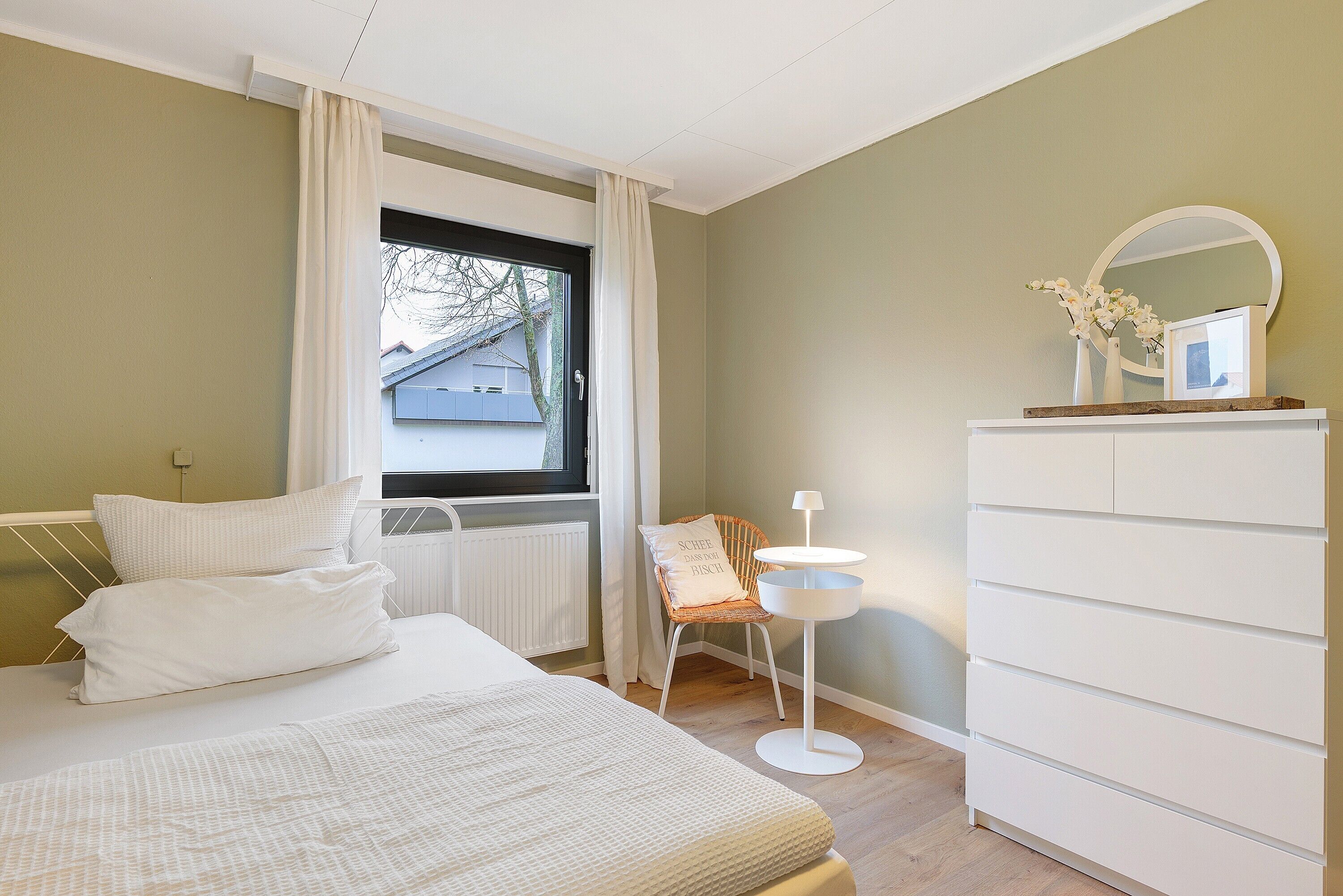 Ein Schlafzimmer mit Bett, Stuhl, Schminktisch und einem Fenster mit Vorhängen sowie einem Spiegel.