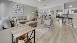 Un vaste espace de vie comprenant un canapé, un pouf et une table à manger. On peut apercevoir au fond une pièce avec un ventilateur de plafond, un lustre et un coin cuisine.
