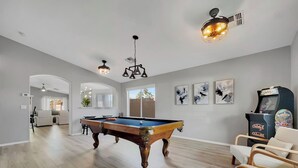 Une pièce spacieuse équipée d’une table de billard, d’un ventilateur de plafond et d’une borne d’arcade rétro.
