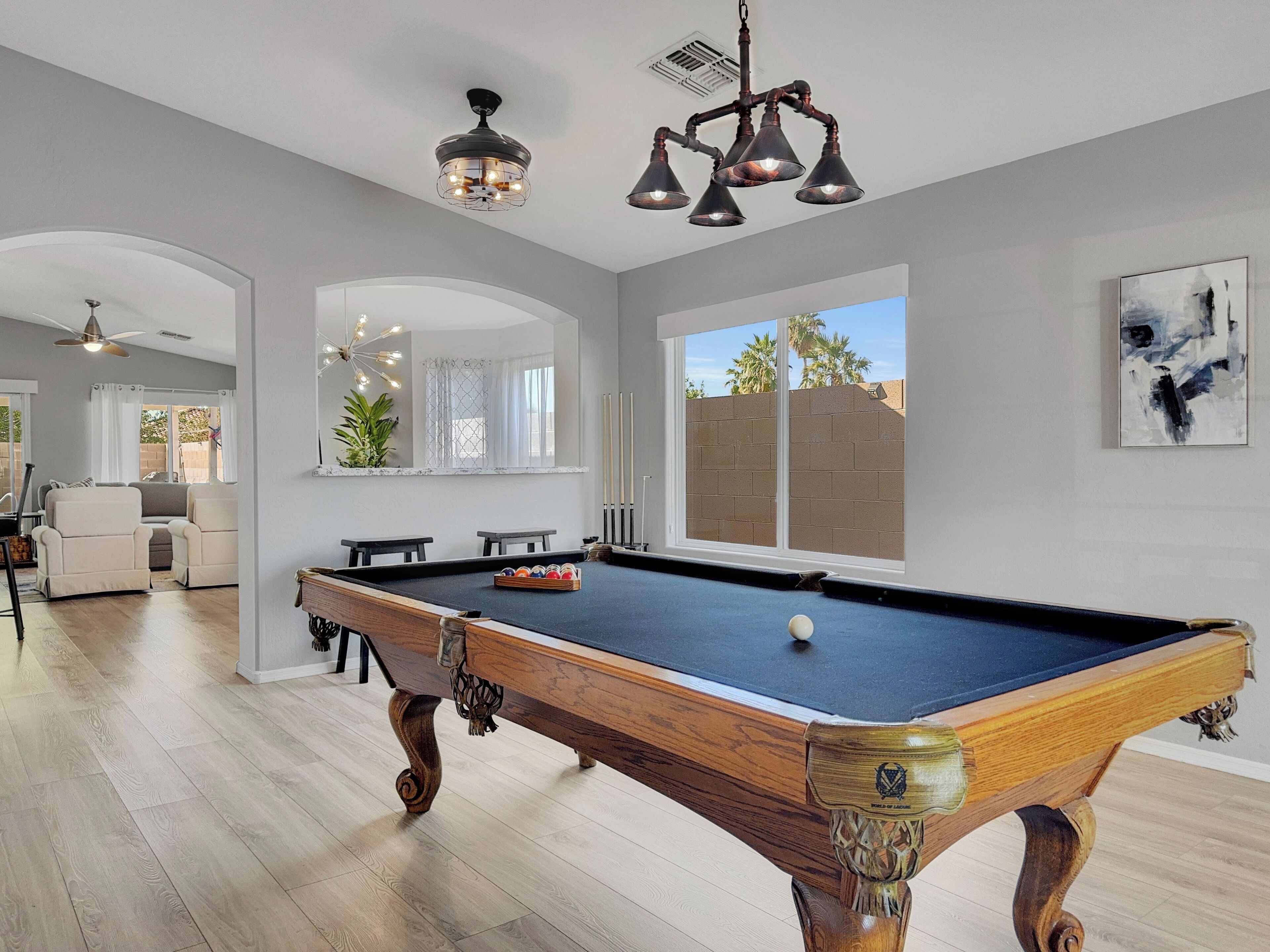 A pool table in a well-lit room with a large window, a ceiling fan, and a modern chandelier.