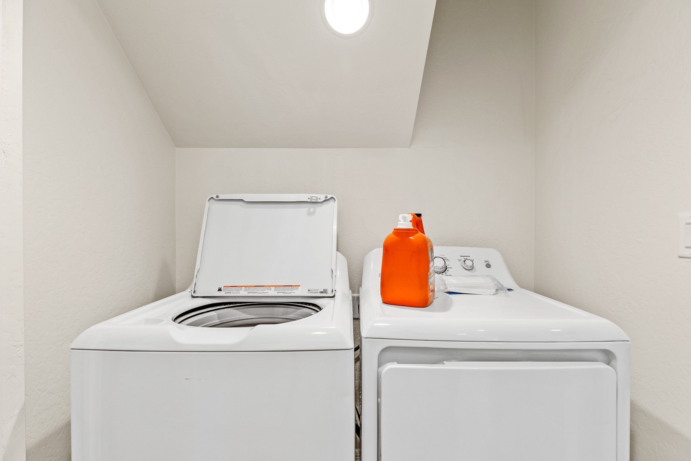 A laundry room with two white washing machines and an orange detergent bottle.