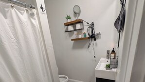 A bathroom with a white shower curtain, a toilet, a sink with a mirror, and shelves with toiletries. - Heart of OTR -Stadiums, Museums, Nightlife, Casino -Walkable Everywhere Cinci (Cincinnati)