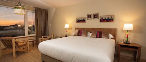 Habitación de hotel con una cama grande, dos mesitas de noche, una silla, una ventana con cortinas y vistas al atardecer.