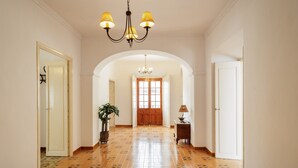 Un couloir avec un sol carrelé, un lustre, une plante en pot et une lampe sur une table d’appoint.