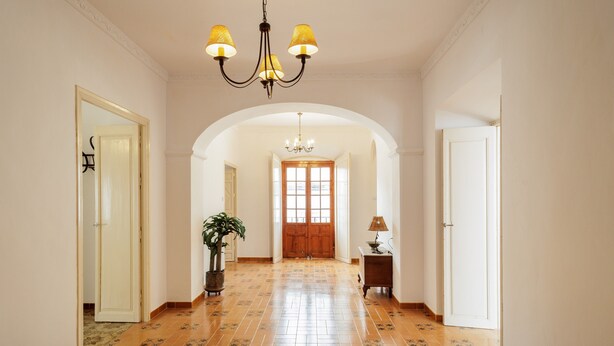 Um corredor com piso de azulejos, um candelabro, um vaso de planta e uma luminária em uma mesa lateral.