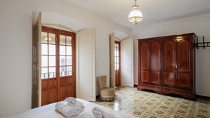 Quarto com uma cama, guarda-roupa de madeira, uma cadeira e uma porta que dá para outro cômodo.
