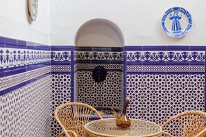 A corner with blue and white patterned tiles, a wicker chair, a small table, and a decorative plate on the wall.