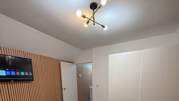 Una habitación de hotel moderna con una cama, un televisor montado en la pared, una lámpara de techo, un sofá y una mesita con un control remoto.