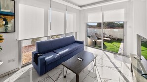 Ein modernes Wohnzimmer mit einem blauen Sofa, einem Glastisch und Blick auf einen Garten mit Gartenmöbeln.