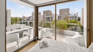 Ein modernes Schlafzimmer mit einem großen Bett, einer Glastür, die auf einen Balkon führt, und Blick auf umliegende Gebäude.