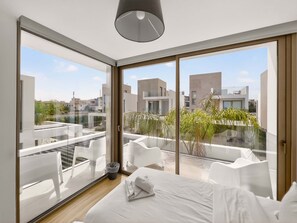 A modern bedroom with a large bed, a glass door leading to a balcony, and a view of surrounding buildings. - Azure Jade Villa | Luxury Retreat, Private Pool (Protaras)