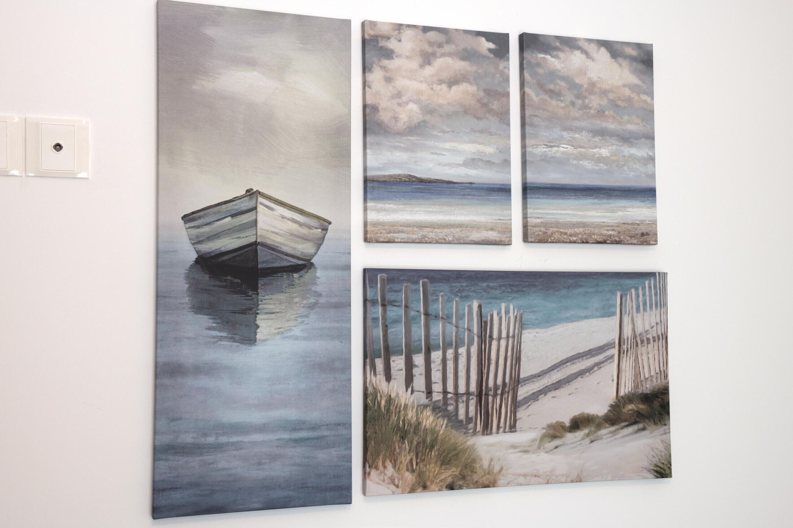 Un mur orné de trois tableaux : un bateau sur l’eau, une plage avec une clôture et un ciel nuageux.