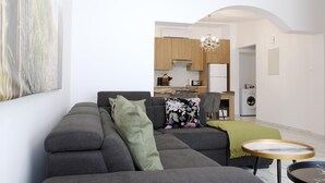 A modern living room with a grey sofa, a round coffee table, and a kitchen in the background with wooden cabinets and a chandelier. - Musan Boutique Inland / Nissi Ave / High-Speed WiFi (Ayia Napa)