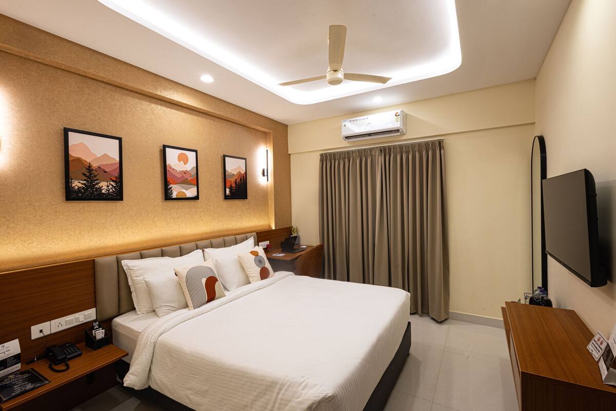 A hotel room with a bed, a desk, a chair, a ceiling fan, an air conditioning unit, and a flat-screen TV.