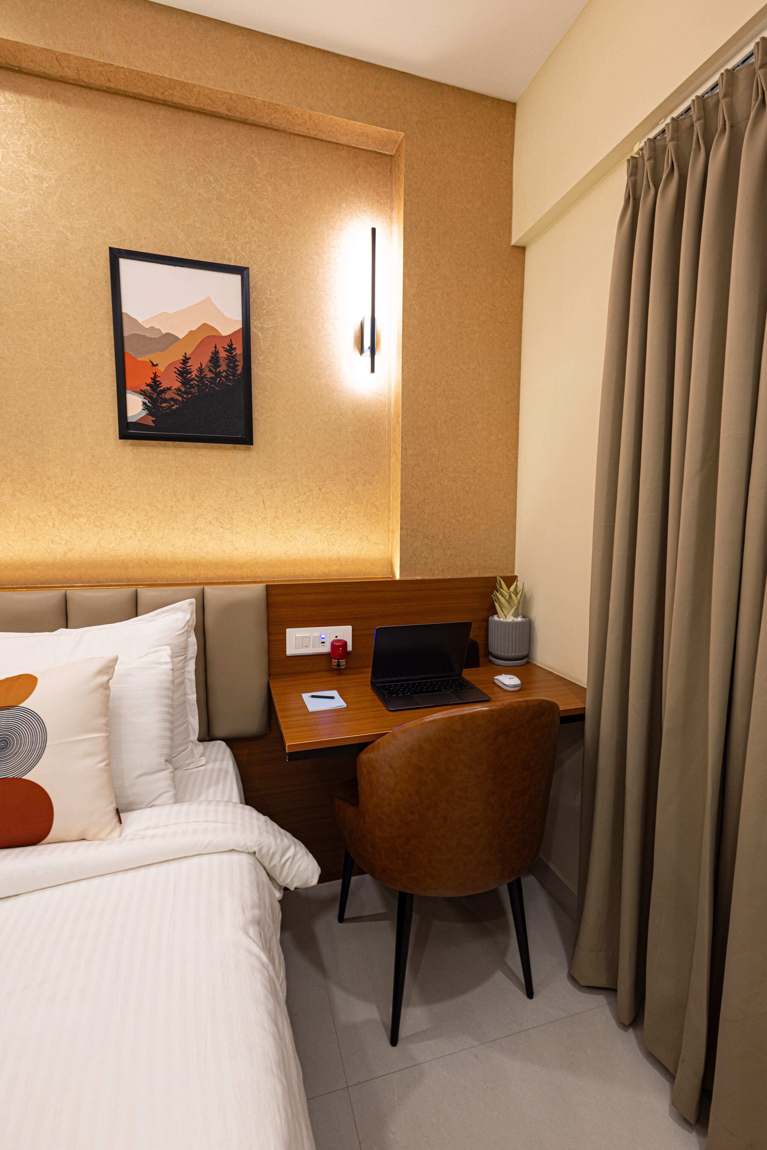 Een hotelkamer met een bed, een bureau met een laptop, een stoel, een plant en een raam met gordijnen.