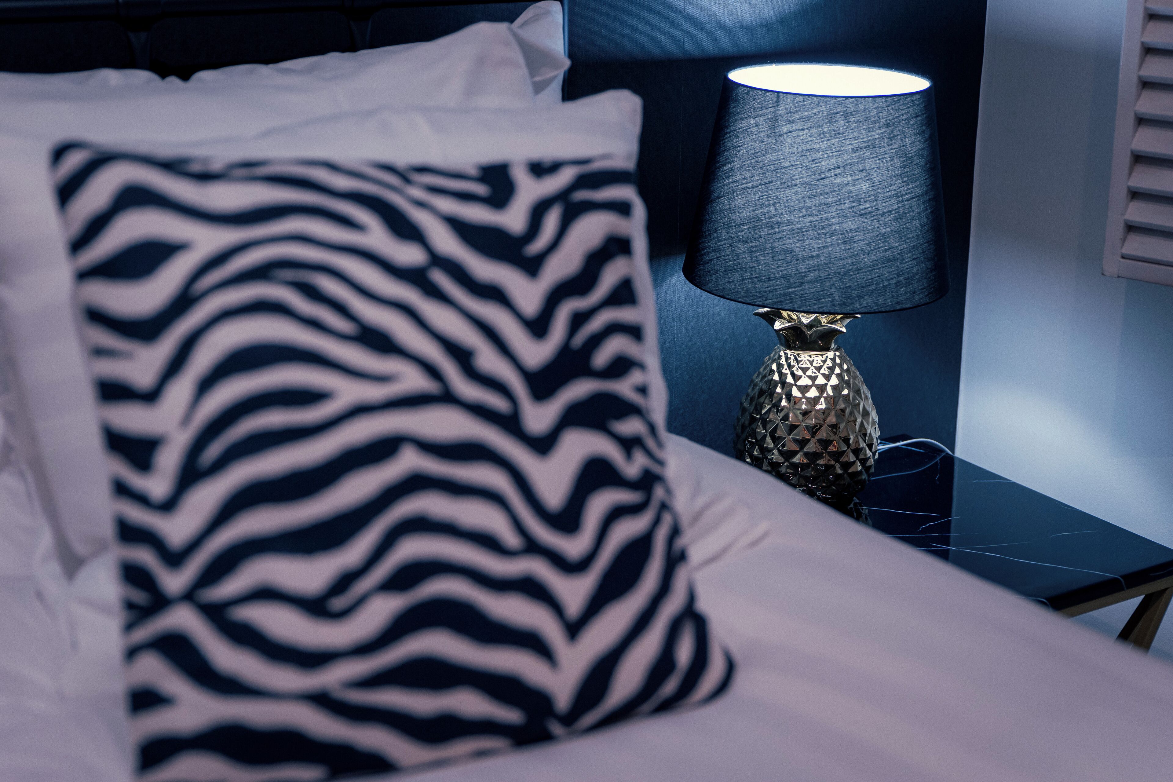 A bed with a zebra-patterned pillow, a lamp with a textured shade, and a bedside table with a geometric-patterned lamp base.