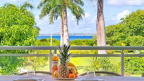 A dining table with wicker chairs, a fruit platter, and a bottle of KARAIBE Punch on a balcony with a view of the sea and palm trees. - La Case à Sucre – Peaceful retreat, sea view, beach front (Anse Marcel)
