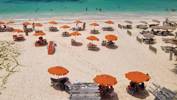 Une plage bordée de rangées de chaises longues et de parasols, une eau turquoise et limpide, et une côte qui s’étend au loin.