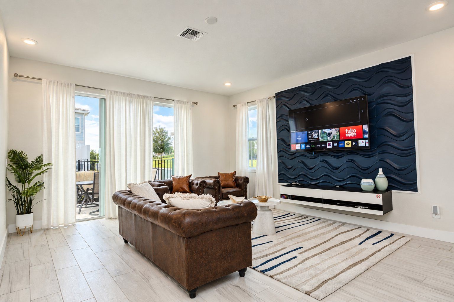 A modern living room with a leather sofa, a flat-screen TV mounted on a textured wall, and a view of the outdoors through large windows.