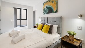 Ein modernes Schlafzimmer mit einem großen Bett, einem grauen gepolsterten Kopfteil, einem Gemälde darüber und einem kleinen Nachttisch mit einer Pflanze.