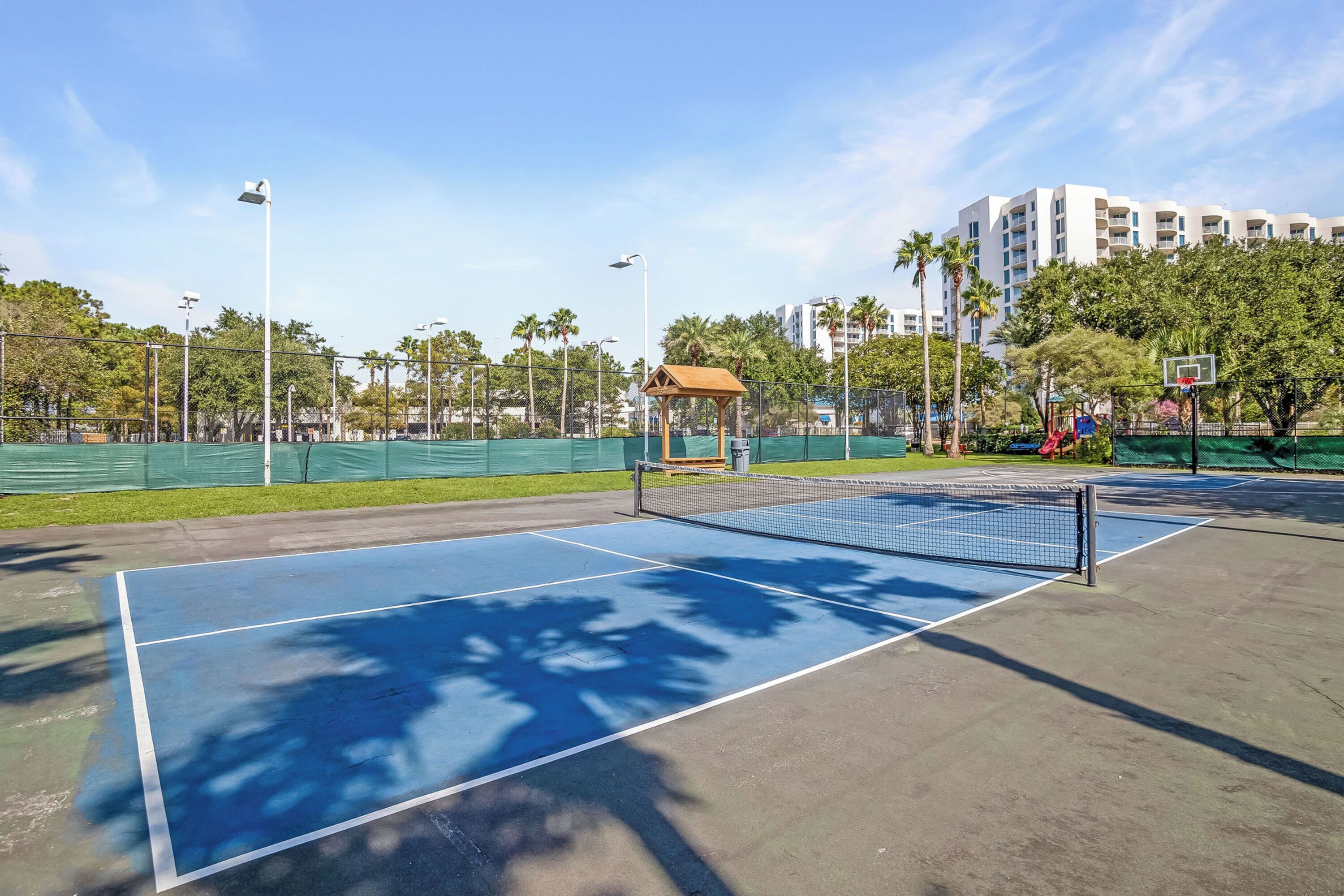 An outdoor tennis court with a net, surrounded by a fence, and a small pavilion.