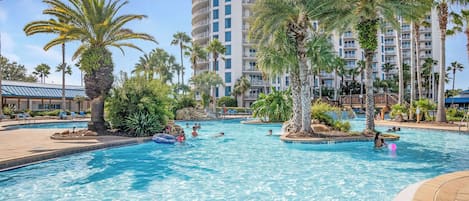 A swimming pool with clear blue water, surrounded by palm trees and a multi-story building with balconies.