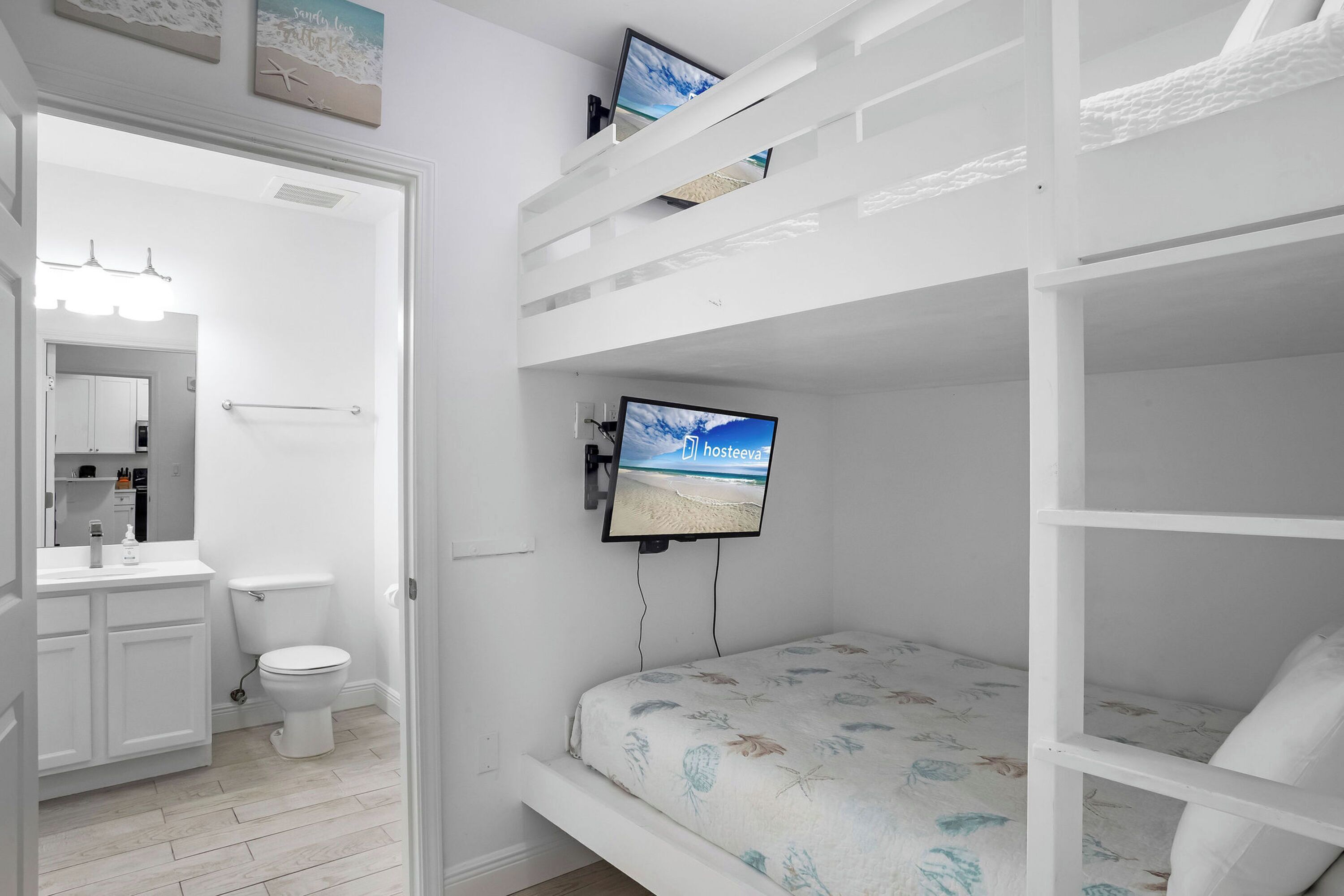 Une chambre avec des lits superposés, un téléviseur fixé au mur, une salle de bain avec des toilettes et un lavabo, et une porte donnant sur une autre pièce.