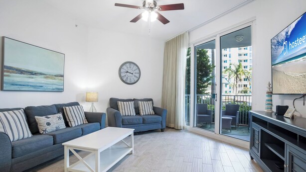 A living room with a sofa set, a coffee table, a wall clock, a ceiling fan, and a TV. - The Palms Destin 2103 (Destin)