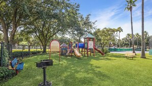 A playground with slides and climbing structures, surrounded by grass and trees. - The Palms Destin 2103 (Destin)