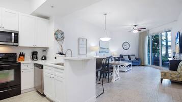 A modern kitchen with white cabinetry, a microwave, and a dishwasher. There is a dining area with a table and chairs, and a living room with a sofa and a ceiling fan.