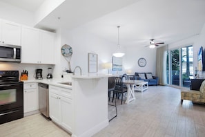 A modern kitchen with white cabinetry, a microwave, and a dishwasher. There is a dining area with a table and chairs, and a living room with a sofa and a ceiling fan. - The Palms Destin 2103 (Destin)