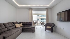 A modern living room with a sectional sofa, a wicker chair, a flat-screen TV, and a large window with curtains. - Apt w/ BBQ & Balcony, 450m From the Beach Sit1202 (Itapema)