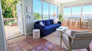 Una habitación con un sofá azul, una mesa redonda pequeña y una silla. Amplias ventanas ofrecen vistas al exterior.