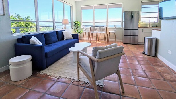 Una sala moderna con un sofá azul, una mesita y una silla. Hay una zona de cocina con un refrigerador y un lavabo. Amplias ventanas ofrecen vistas al exterior.