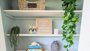 Interior de una vivienda con una escalera que conduce a una plataforma. La plataforma tiene tres repisas con varios objetos: una cesta de mimbre, un letrero de bienvenida y una planta colgante.