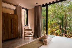 Quarto com cama, guarda-roupa de madeira, uma cadeira e uma janela ampla com vista para o verde.