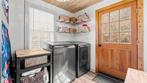 Eine kompakte Küche mit zwei Kühlschränken, Holzregalen darüber, einer Holztür und einem Fenster mit Jalousien.