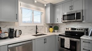 A modern kitchen with grey cabinets, a stainless steel dishwasher, a built-in oven, a microwave, and a coffee maker. - Newly Renovated 3BR in Kingston’s Desirable West End | The Yellow Door Retreat (Kingston)