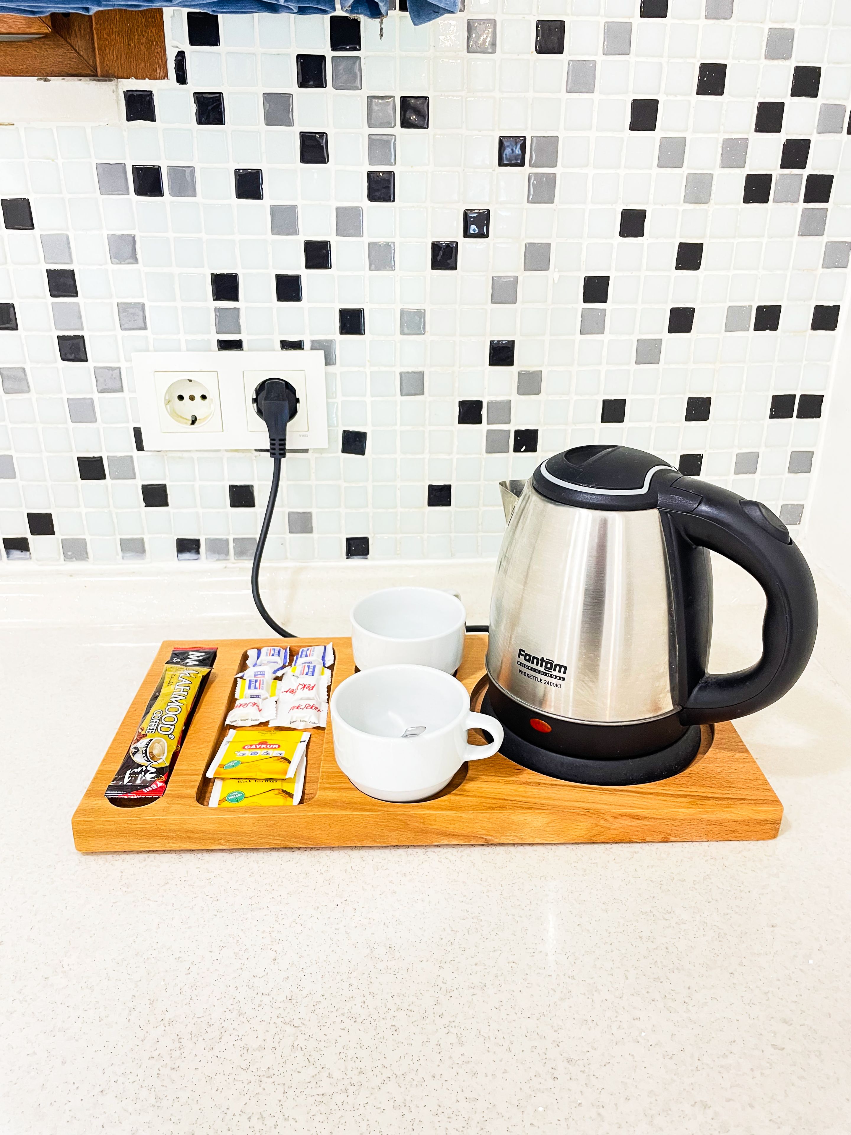 A kitchen counter with a coffee maker, two white cups, a wooden tray with coffee pods and a chocolate bar, and a tiled backsplash.