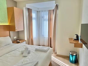 Quarto de hotel com cama bem arrumada, mesa de cabeceira com controle remoto, uma pequena planta, garrafa de água, chaleira e lixeira.