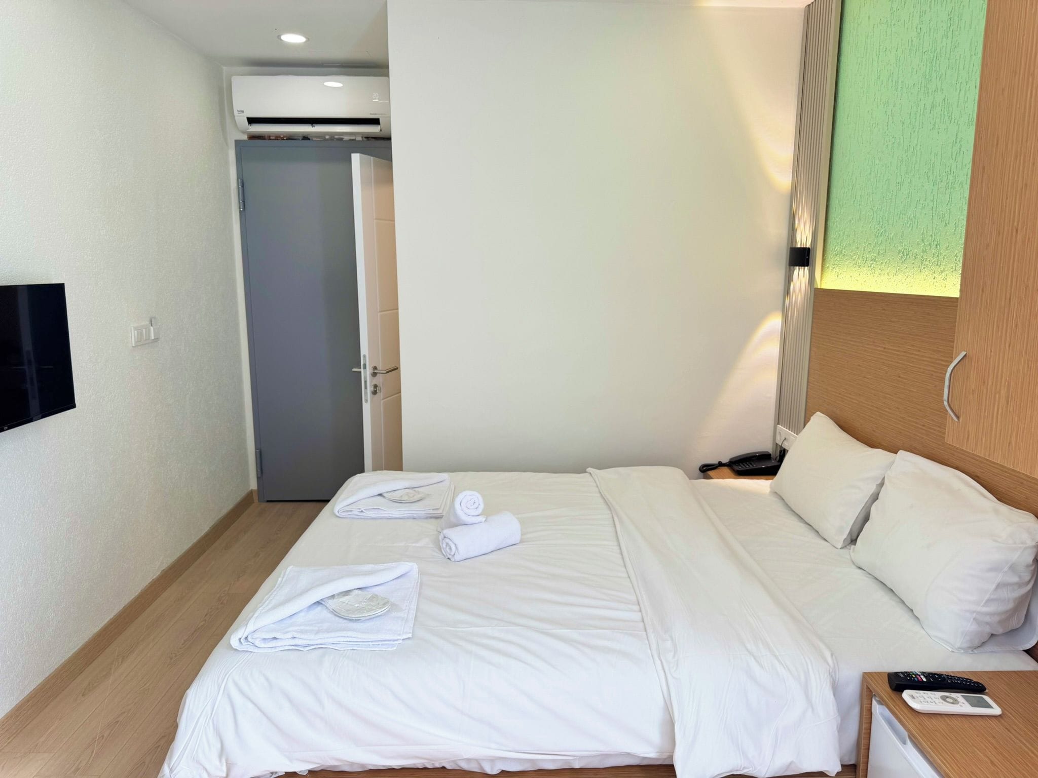 A hotel room with a bed, a bedside table with a phone and a remote, a wall-mounted TV, and a door leading to another room.