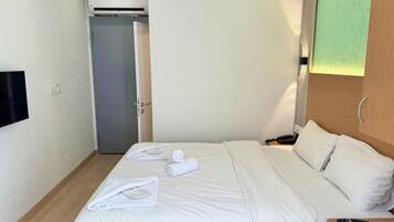 A hotel room with a bed, a bedside table with a phone and a remote, a wall-mounted TV, and a door leading to another room.