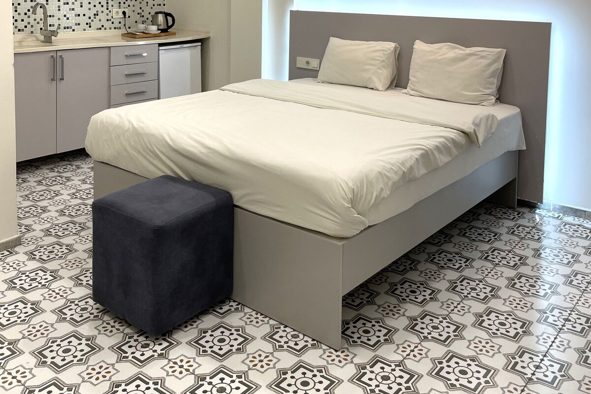 Quarto com cama, mesa de cabeceira, um banquinho, janela com cortinas, ar condicionado, pequeno espaço de cozinha com pia, cafeteira e ladrilhos no chão estampados.
