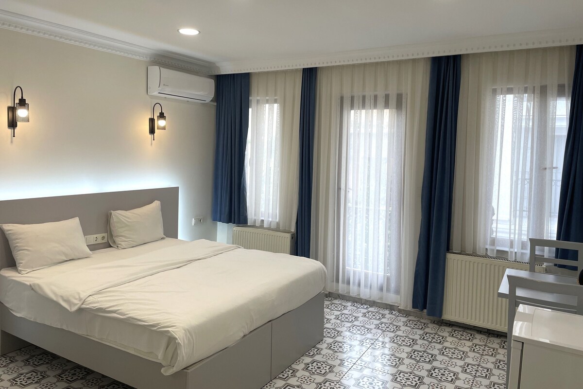 Quarto de hotel com uma cama, escrivaninha, radiador e janela com cortinas.