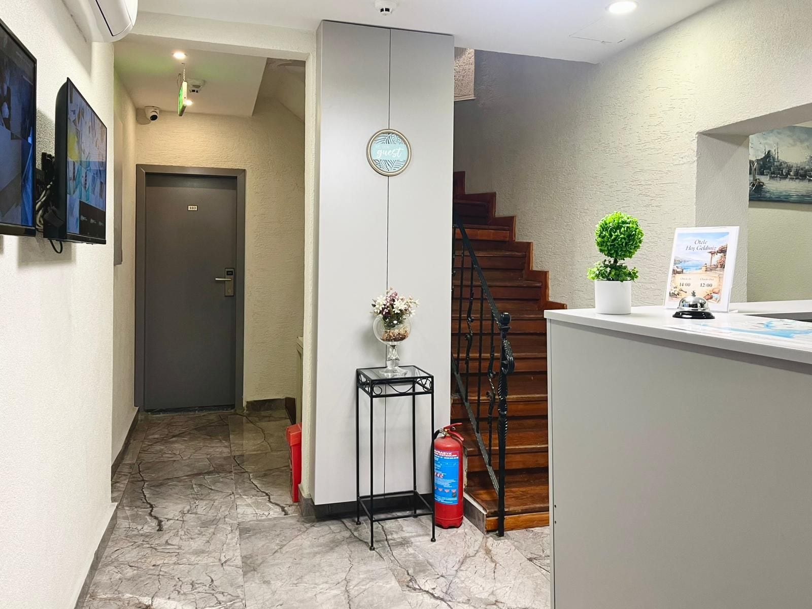 A hotel lobby with a reception desk, a clock, a fire extinguisher, a staircase, and a door.