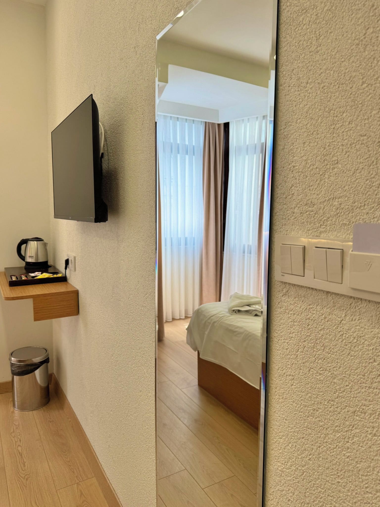 A hotel room with a flat-screen TV mounted on the wall, a wooden bedside table with a kettle, a small trash bin, and a bed with a white duvet.